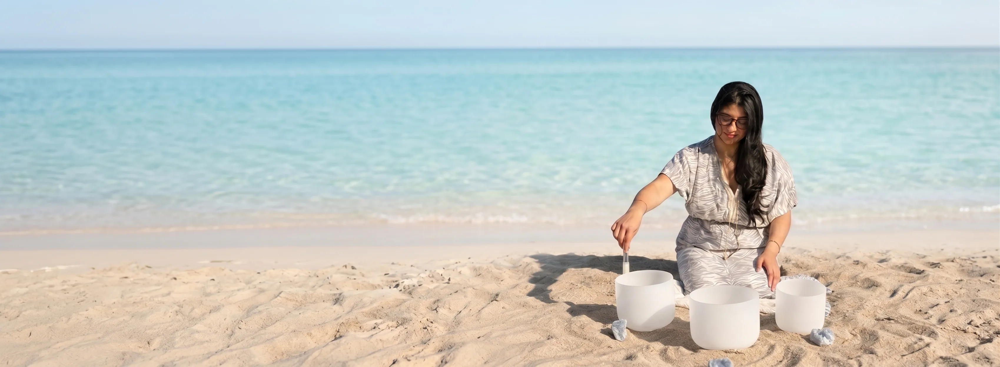 Sylvia Lee kneeling on the beach playing crystal singing bowls by the sea